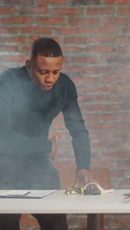 doubting african-american scientist disperses smoke of burning equipment during failed experiment at table with laptop in laboratory