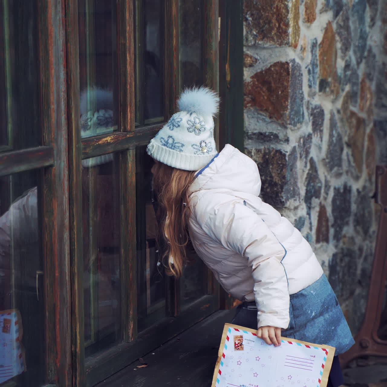 Little girl is looking through the small wooden windows outdoors. Curious child looks inside the house with fairy atmosphere while standing near the windows in winter.