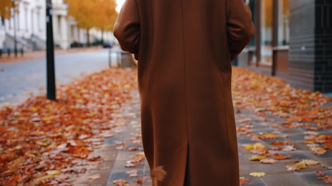 Person i brun kåpe som går på et fortau dekket av høstblader