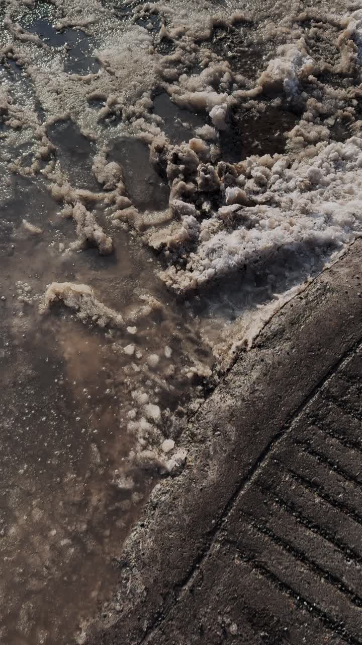 POV di un pedone con scarpe da ginnastica che attraversa una strada urbana in mezzo a fanghiglia sporca e acqua di fusione sul bordo del marciapiede a marzo. Condizioni realistiche di camminata all'inizio della primavera per video social e uso editoriale