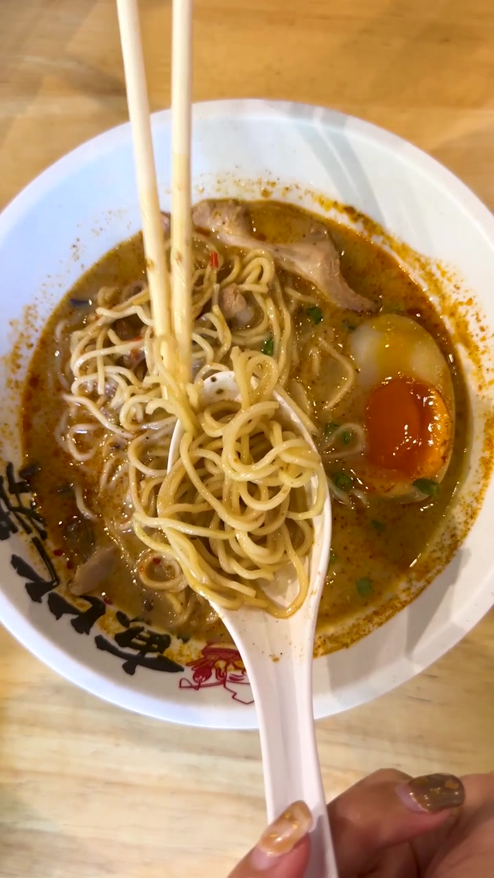 Una mano usa bacchette e cucchiaio per sollevare i noodles ramen in un ambiente di ristorante giapponese ben illuminato
