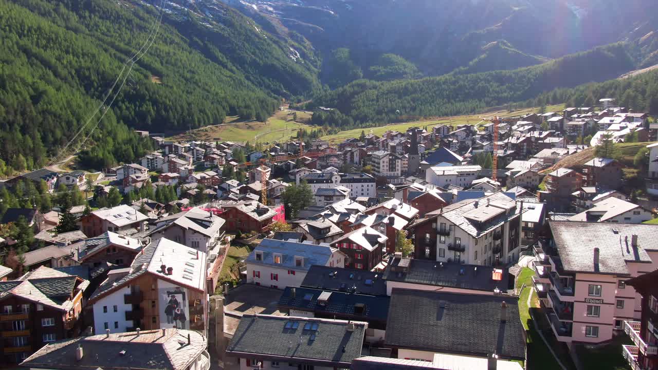 Renkli dağ evleri ve yeşil çayırlarla dolu canlı Alp kasabası Saas-Fee'den heybetli karlı dağlara doğru havadan yukarı eğimli çekim.