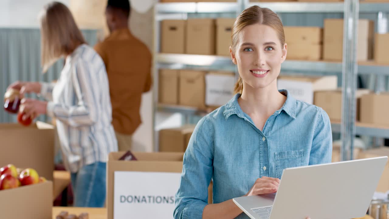 Close-up view of caucasian young female volunteer typing on laptop and smiling to the camera while her coworkers packing donation boxes in charity warehouse