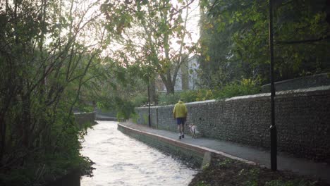 man in yellow jacket walks alongside river with playful dog.