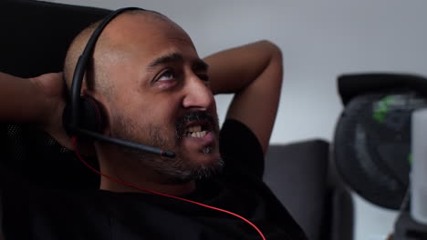 hombre calvo asiático girando en una silla en casa hablando usando auriculares con el brazo detrás de la cabeza