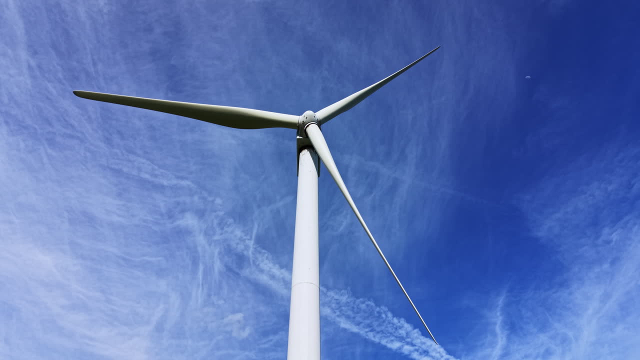 Turbina eolica si erge alta contro un cielo azzurro limpido con nuvole leggere di giorno, mostrando l'energia pulita in azione.