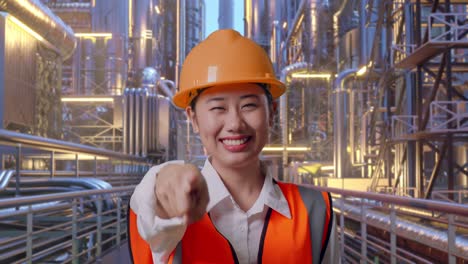 Close Up Of Asian Female Engineer With Safety Helmet Smiling And Touching Her Chest Then Pointing At You While Standing At A Vast Oil Refinery