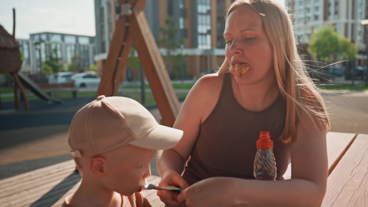 Kvinde og ung dreng sidder sammen på en parkbænk og nyder tiden udendørs. Den voksne holder barnets hånd, mens begge interagerer med små genstande i deres hænder. I nærheden er en plastikflaske med drikke. Sollys