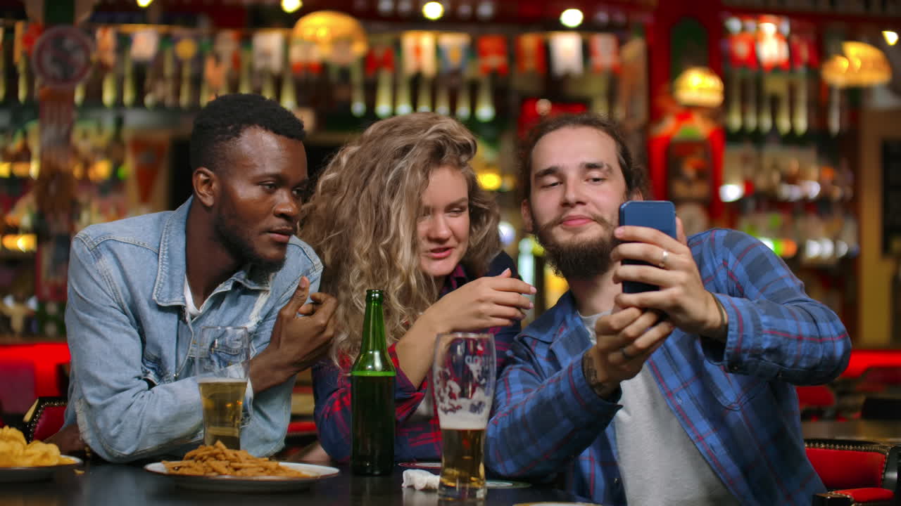 un gruppo multietnico di amici si fa un selfie in un bar e ride con una birra mentre guarda le foto sullo schermo di uno smartphone