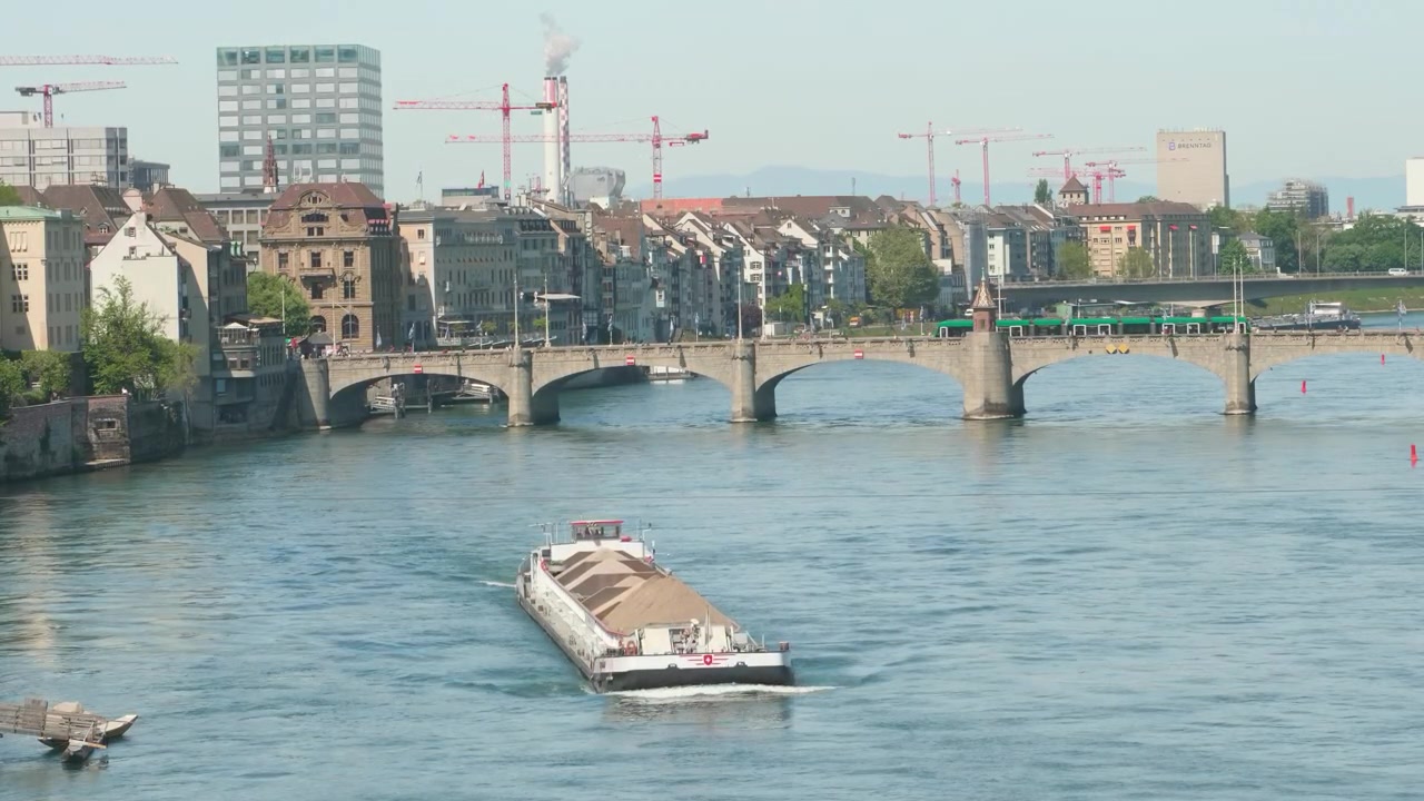 Inquadratura statica in una giornata di sole velata, una chiatta da carico piena naviga il fiume Reno verso il ponte Mittlere Brücke, skyline di Basilea con gru sullo sfondo.