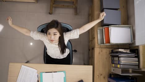 view from above smiling asian businesswoman is stretching her body and looking up into distance with folded arms while taking a break from work at workplace.