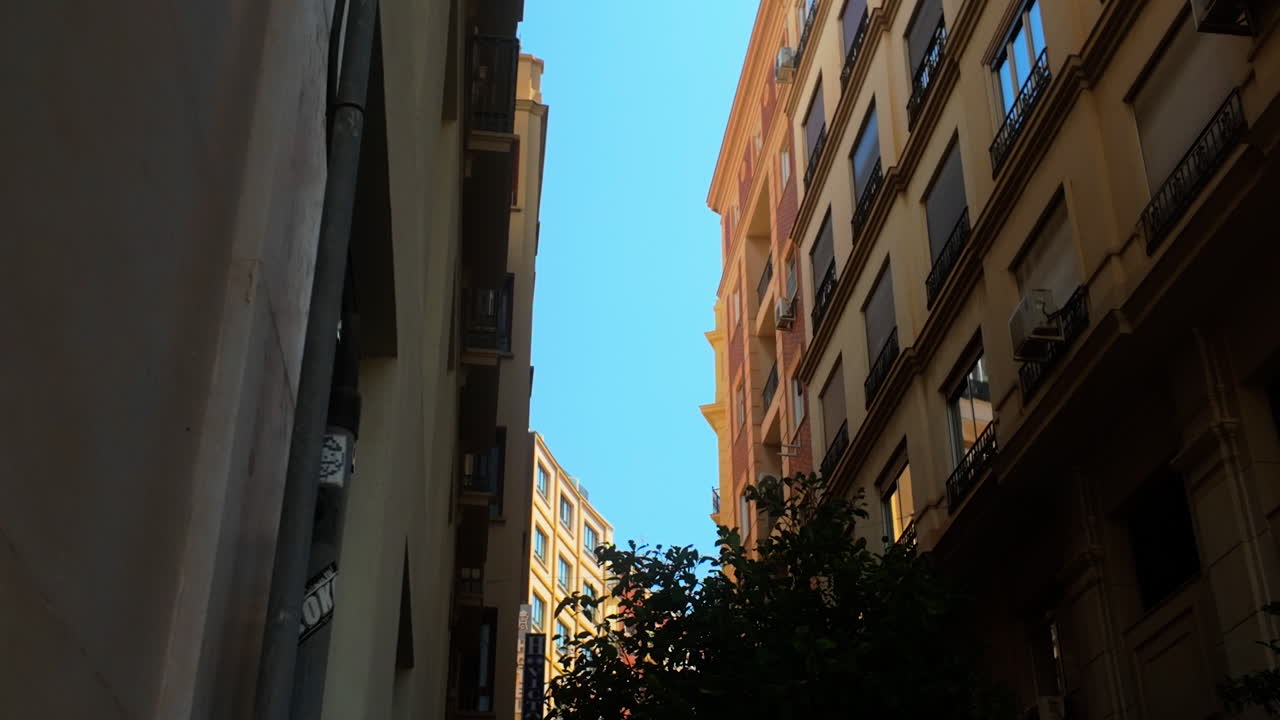 Looking up through a narrow street in Málaga, Spain, surrounded by Mediterranean-style buildings with balconies, showcasing the vibrant urban architecture under a clear blue sky