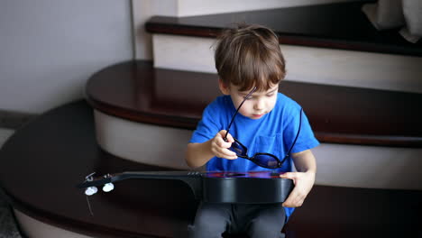 lovely male toddler sitting on the stairs with guitar in his hands. kid puts his sunglasses beside him. top view.