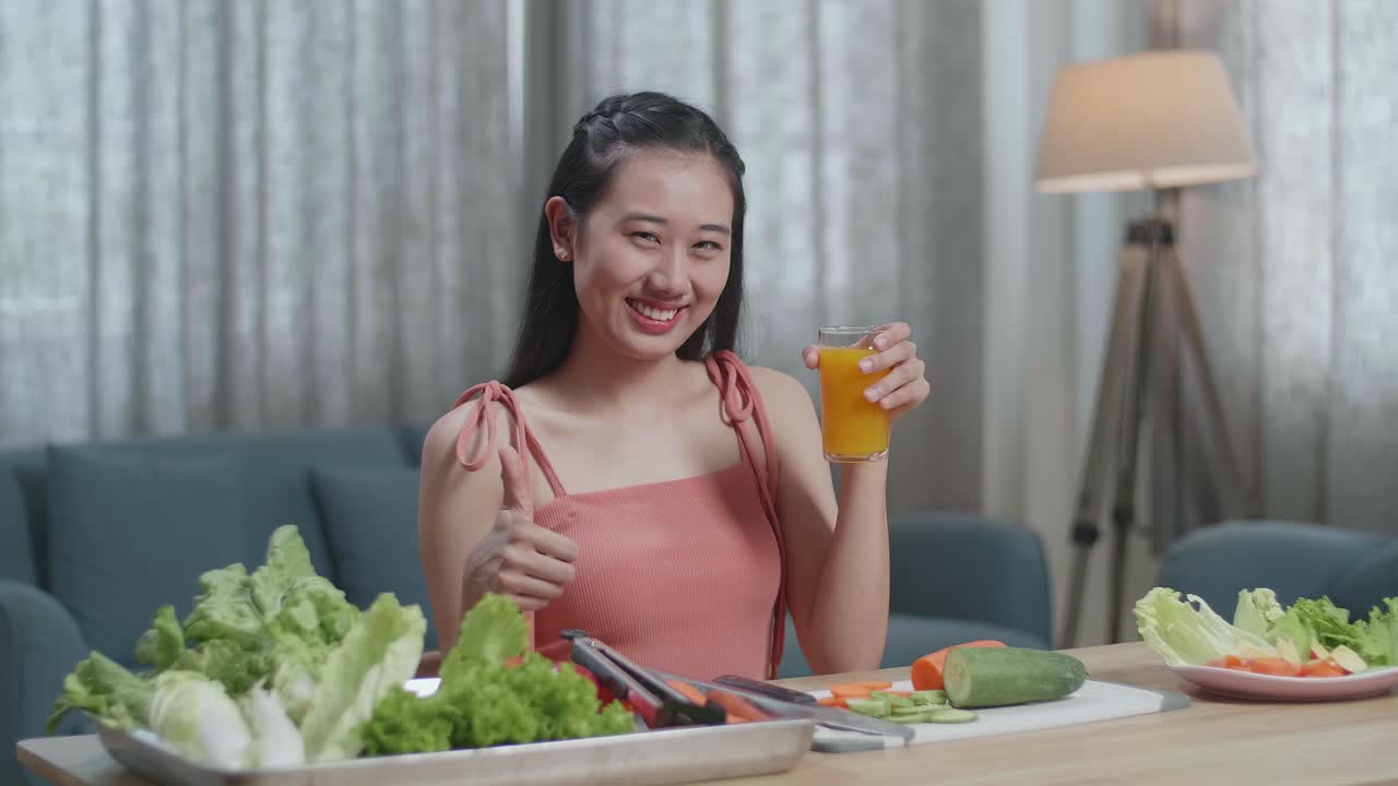 행복 한 아시아 여성 이 오렌지 주스 한 잔 을 들고 집 에서 건강 한 음식 을 먹는 동안 엄지손가락 을 올려 놓은 몸짓 을 하고 있다