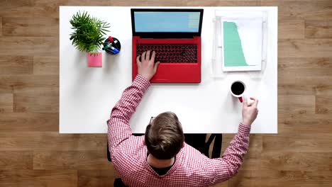 young man in glasses typing on laptop and drinking coffee, topshot, sitting behind table with documents