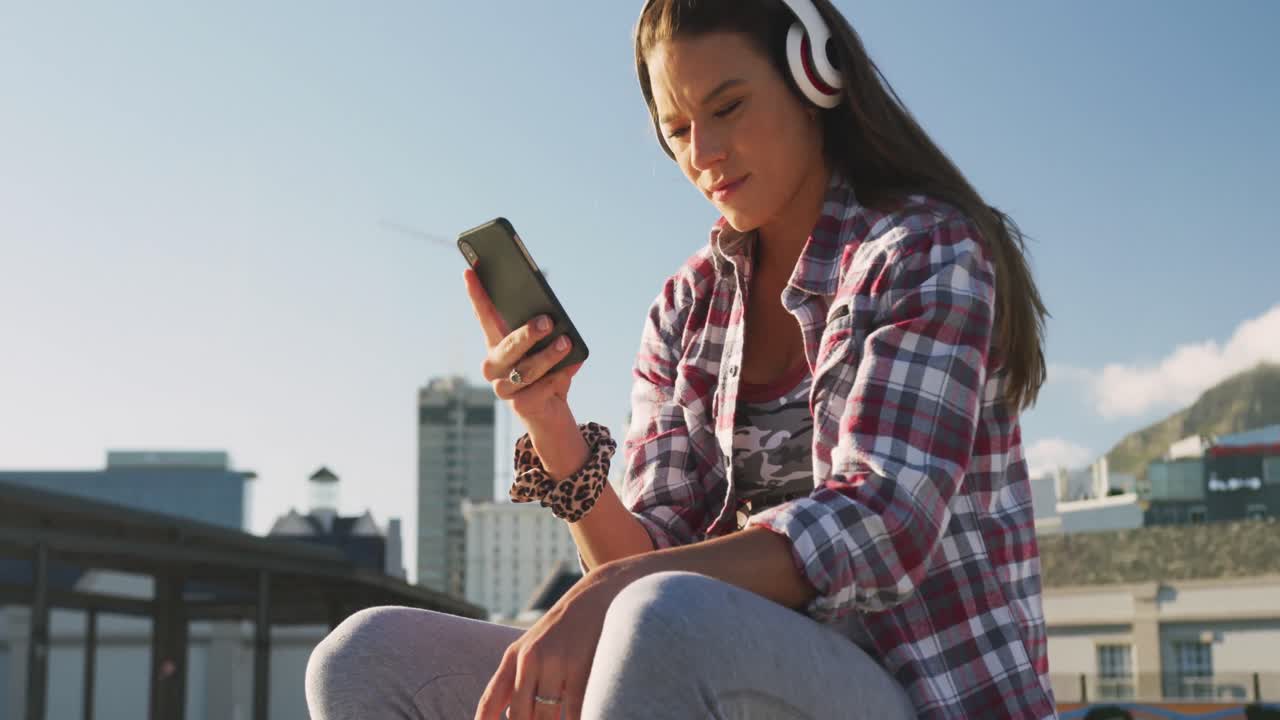 mujer caucásica sonriente con auriculares y usando un teléfono inteligente en un skatepark