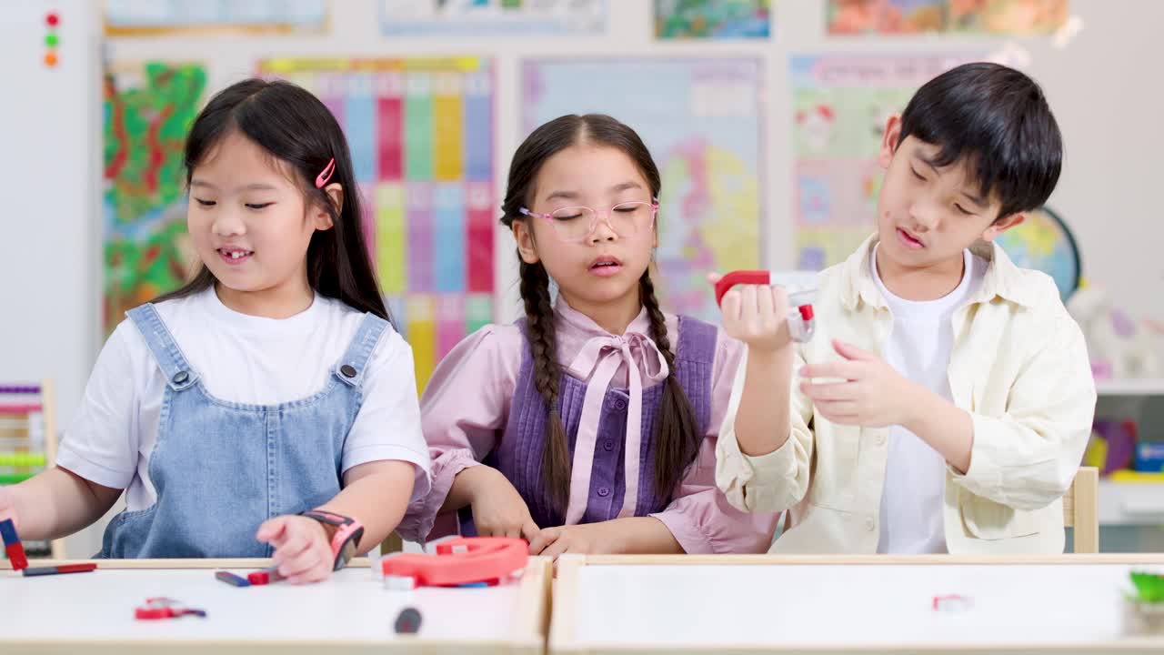 Three Asian primary students explore magnets together in bright classroom, static camera, cheerful educational mood