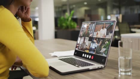 mujer biracial usando una computadora portátil para una llamada de video, con diversos colegas de negocios en la pantalla