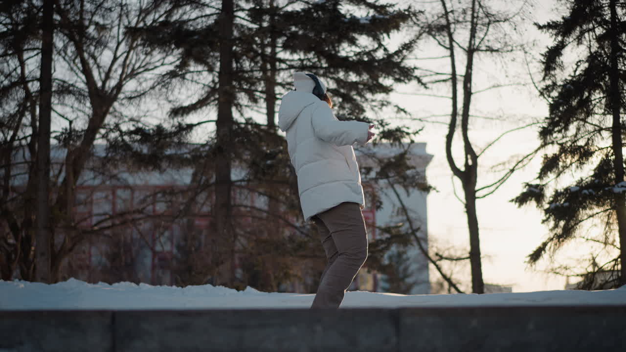 Performer standing on step pavement near snow swaying body to music rhythm wearing white puffer jacket beanie and headphones under warm sunset light against blurred forest backdrop