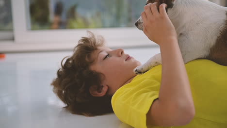 side view of a blond boy with curly hair lying on the floor and caresses his dog