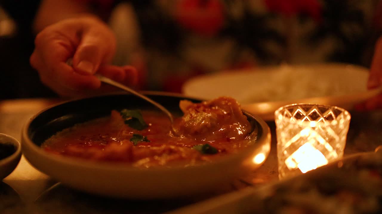 A person serves curry by candlelight, creating a warm, intimate dining atmosphere. The setting is cozy and inviting