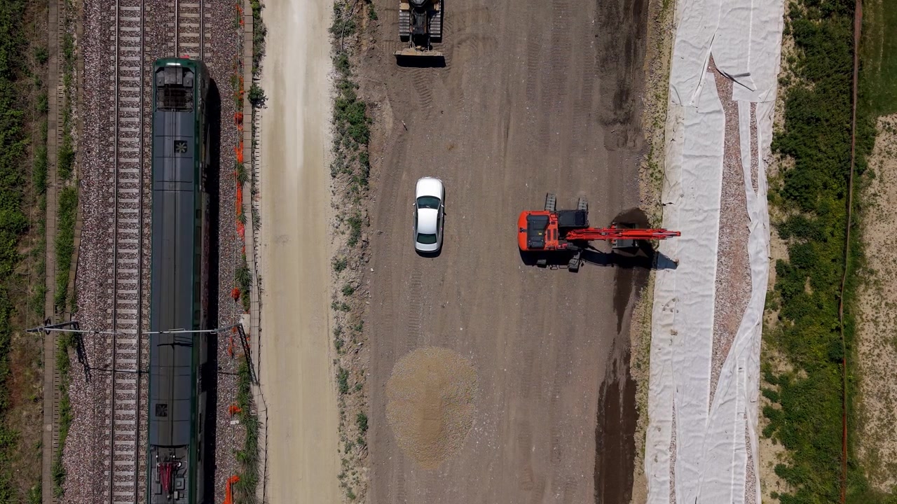 Railway construction site in northern Italy. An excavator levels soil beside active rail tracks while a passing train highlights transport infrastructure expansion and engineering work.