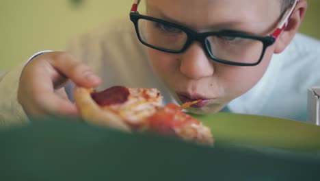 cute boy in glasses has lunch with pizza at table closeup