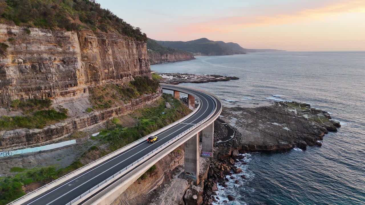 Stunning drone footage of the Sea Cliff Bridge, revealing a car traveling along the winding road with panoramic ocean views
