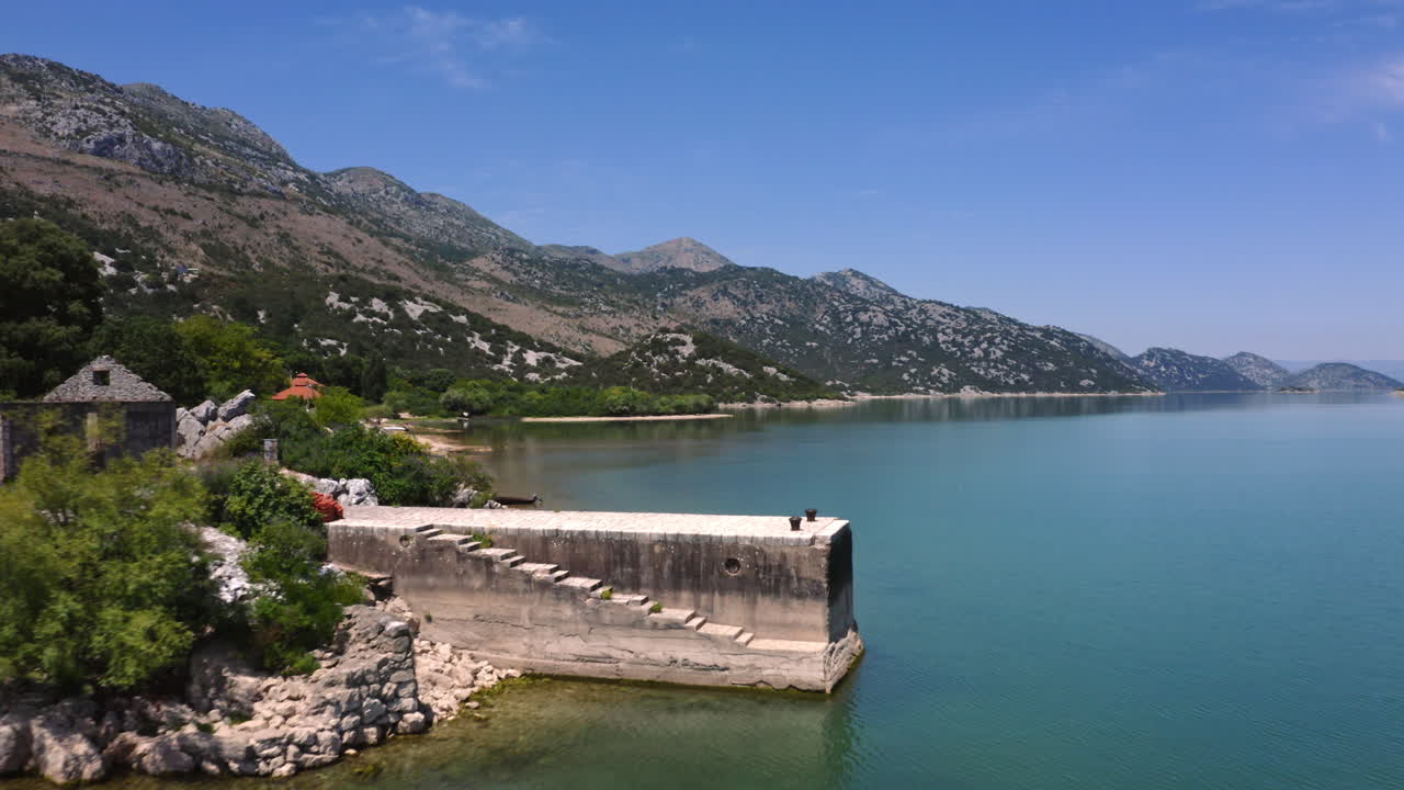 The picturesque shoreline of lake Skadar in Montenegro with tall mountains looming above, trees, stony beaches and cottages around and a stone pier emerging from the coast, aerial scenic view 4K.