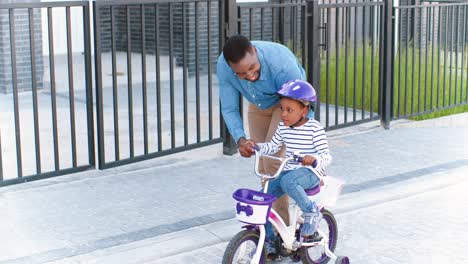 african american father teaching small girl in helmet riding on bike at street in suburb