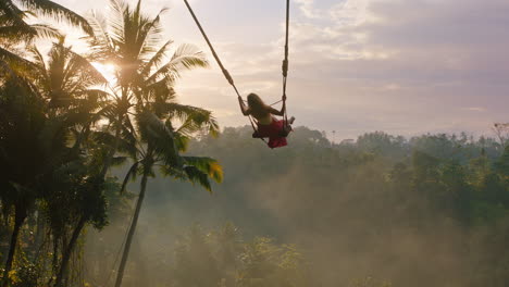 Eine-Reisende-Frau-Schwingt-Sich-Bei-Sonnenaufgang-über-Den-Dschungel-Und-Genießt-Einen-Exotischen-Urlaub-Auf-Einer-Schaukel-Sitzend,-Während-Die-Sonne-Durch-Palmen-Im-Tropischen-Regenwald-Scheint.-Urlaubs-Lifestyle-Freiheit