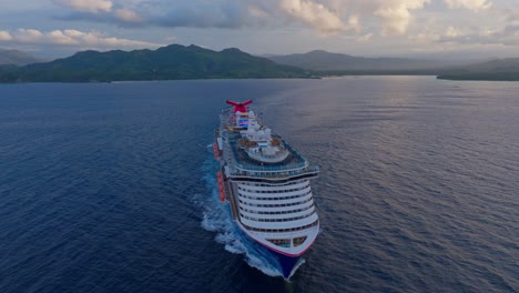 aerial orbit shot of cruising cruise ship with luxury pools and tv monitor on rooftop in dominican republic - sunset time and clouds at sky