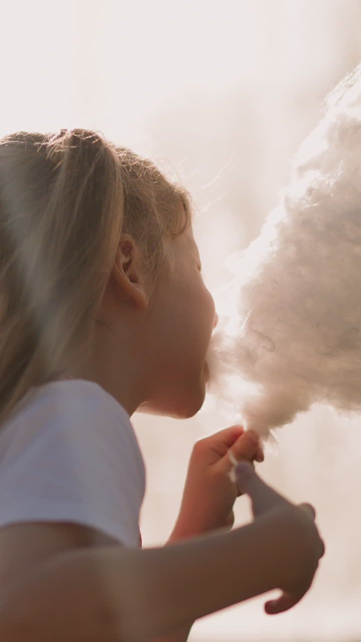 Pretty girl eats sugar cotton wool against blurry fountain closeup slow motion. Preschooler blonde child enjoys dessert. Happy time in amusement park