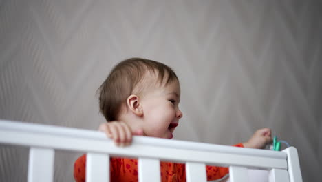 cheerful happy baby boy standing in the cot waving hand with pacifier. adorable kid in orange shirt smiling sweetly in his crib.