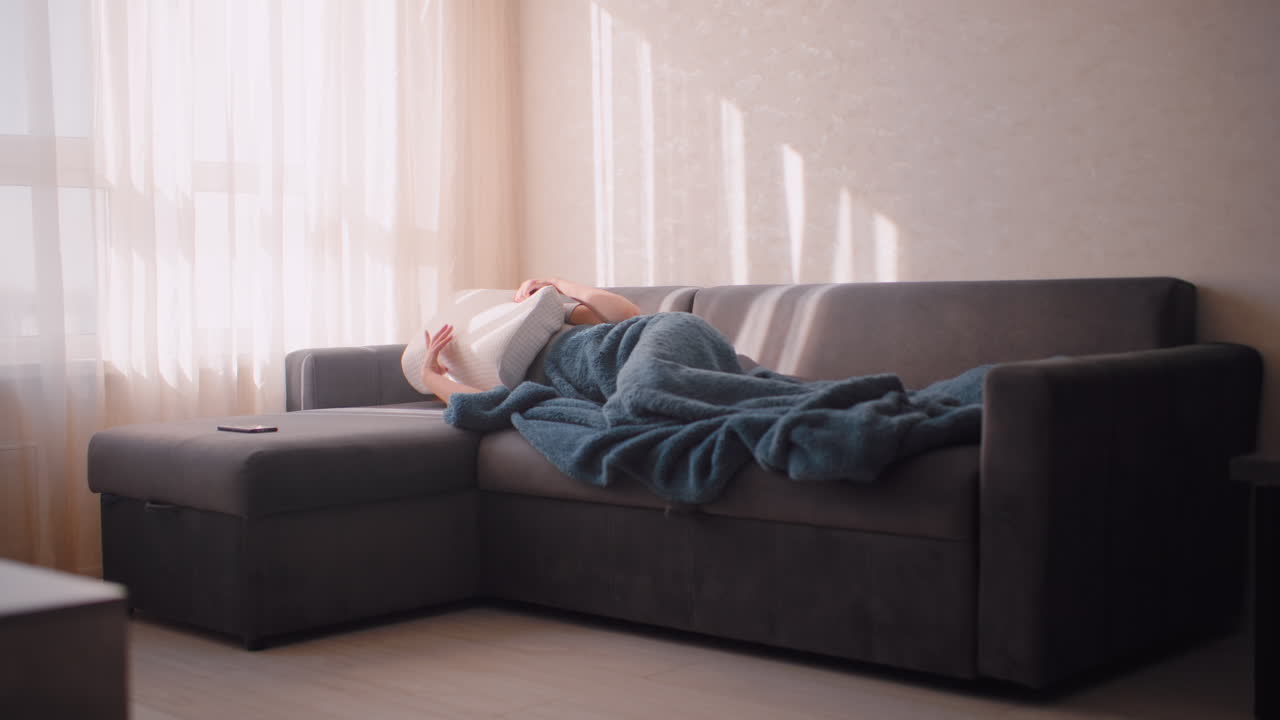 Young woman lying on dark sofa wrapped in blue blanket covering face with pillow to block out strong sun rays beaming through curtain as nearby phone rests on couch in peaceful quiet room