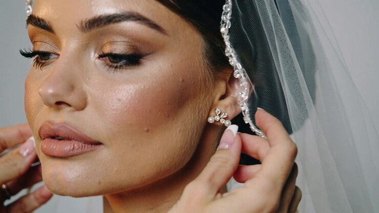 Le mani dello stilista si protendono per sistemare il bordo di perline della sposa, fissando un orecchino di perla per la cerimonia in studio.