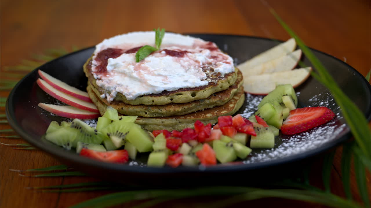 Ripresa in slow motion con allontanamento di un piatto con pancake sani di avena verde e matcha, serviti con fette di mela, kiwi e fragola e panna montata