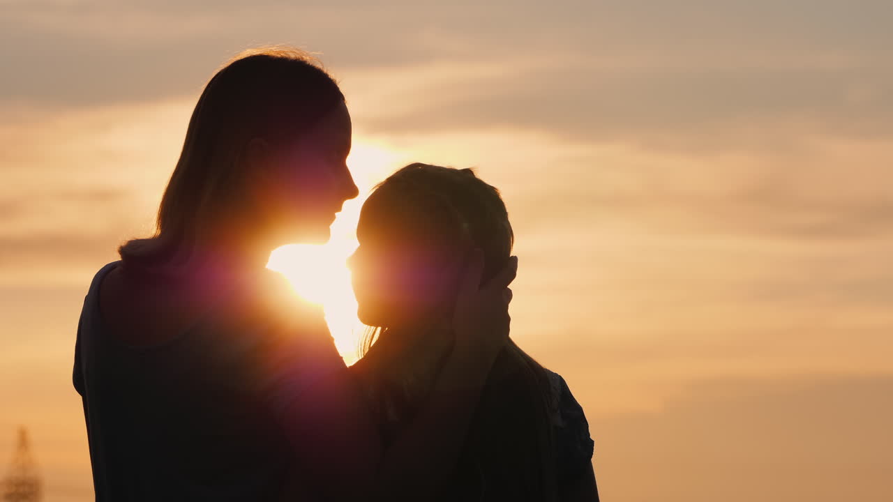 una mujer besa suavemente las siluetas de su hija contra el cielo donde se pone el sol
