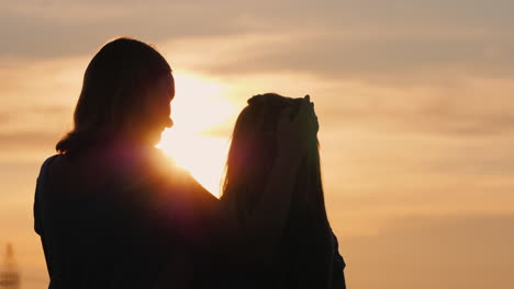 a woman gently kisses her daughter silhouettes against the sky where the sun sets