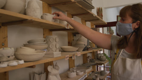 Female-caucasian-potter-wearing-face-mask-and-apron-using-digital-tablet-and-checking-pottery-stock-