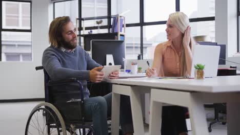 Disabled-caucasian-businessman-sitting-on-wheelchair-using-digital-tablet-talking-to-coworker-in-off