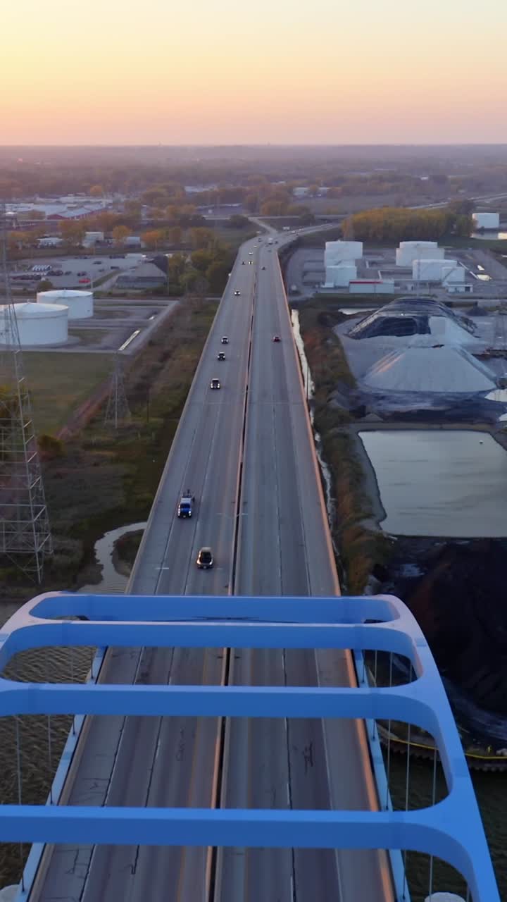 Cars move along a tall blue arch bridge stretching over a river as the sun sets beyond the city, illuminating industrial buildings and autumn trees in soft golden light