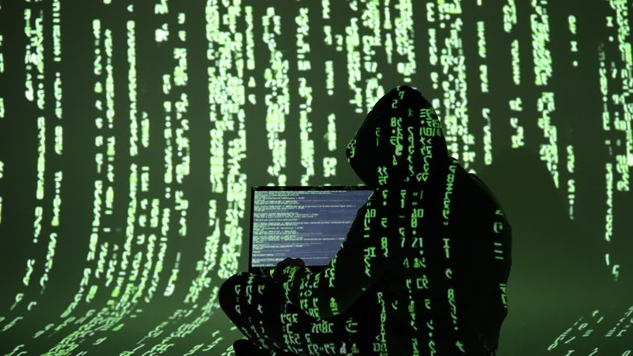 A cybercriminal in a hooded outfit types on a laptop surrounded by digital green code. This immersive matrix-style scene reflects cybercrime, hacking, and data breaches in progress