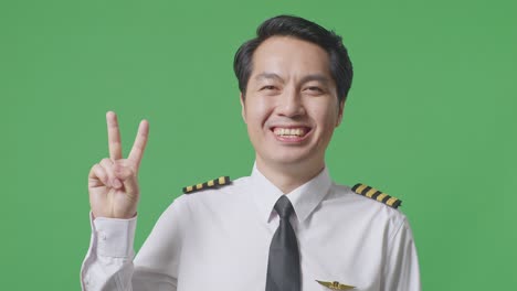 녹색 스크린 배경 스튜디오에 서있는 동안 카메라에 평화의 몸짓을 보여주고 미소 짓는 아시아 남자 조종사의 클로즈업