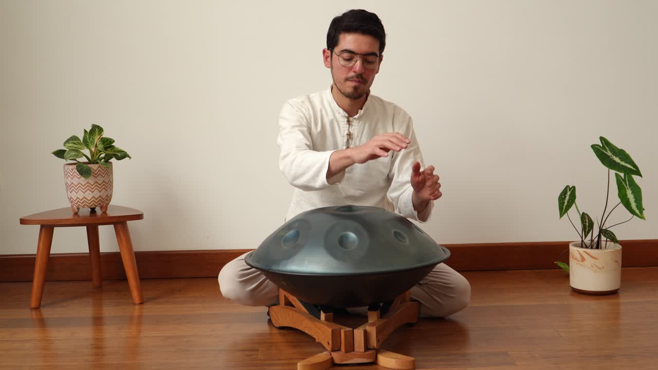 Front-view of a man seated cross-legged, playing melodies on a handpan (hang drum) in a calm minimalist studio with wooden floor and plants—ideal for wellness, ambient music and meditation themes