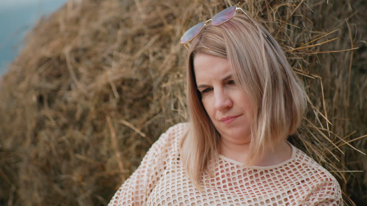 женщина, опирающаяся на сено, носящая солнцезащитные очки на голове и сетчатую ткань, вдумчиво открывая новую страницу в книге, спокойная сельская местность с теплым солнечным светом