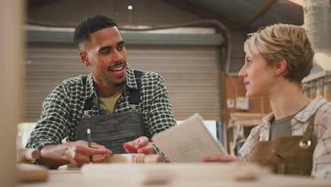 Male-And-Female-Apprentices-Working-As-Carpenters-In-Furniture-Workshop-Measure-Wood-And-Take-Notes