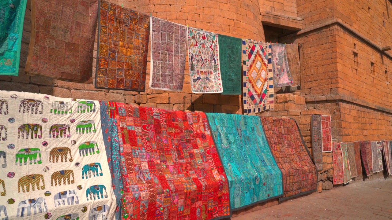 Cinematic static shot of vibrant Rajasthani embroidered tapestries and patchwork textiles hanging on yellow sandstone walls in Jaisalmer, Rajasthan, India.