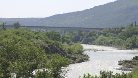 cars driving on the bridge spanning the river at daylight with mountains views in the background