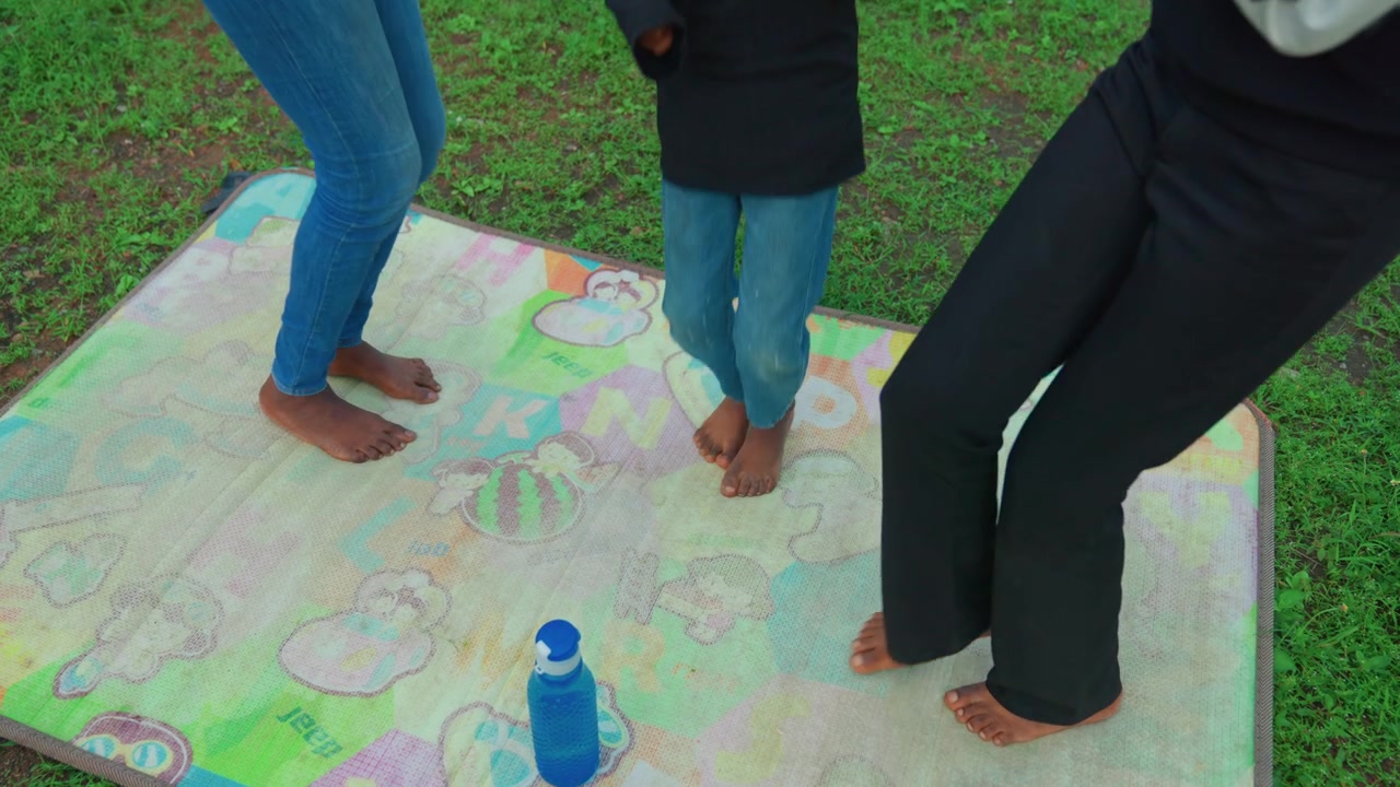 Barefoot black mother and child dancing on patterned mat in green lawn, playful stomps, water bottle marker, denim outfits, upbeat mood, joyful family connection, spontaneous summer activity
